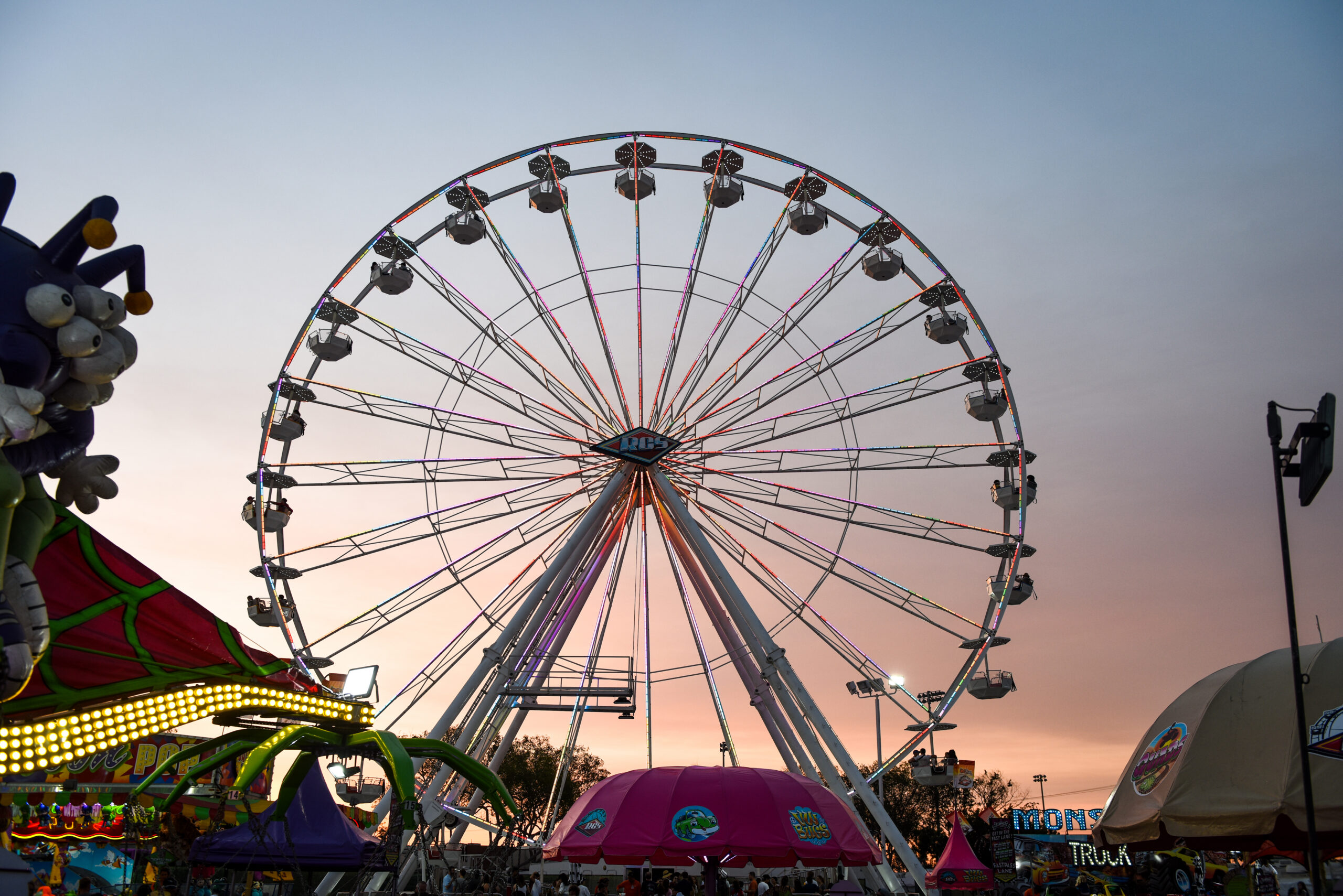 A large Ferris wheel is lit up against a colorful sunset sky at the OC Fair Costa Mesa, with tents, rides, and booths visible in the foreground.