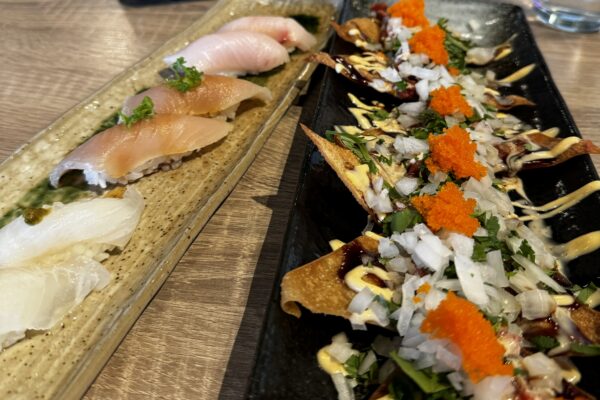 A close-up of two stylish plates at Yuzu Sushi Costa Mesa: one with assorted sushi pieces, and another with crispy nachos topped with diced onions, herbs, sauces, and bright orange roe. Bowls of soy sauce and pickled ginger are in the background.