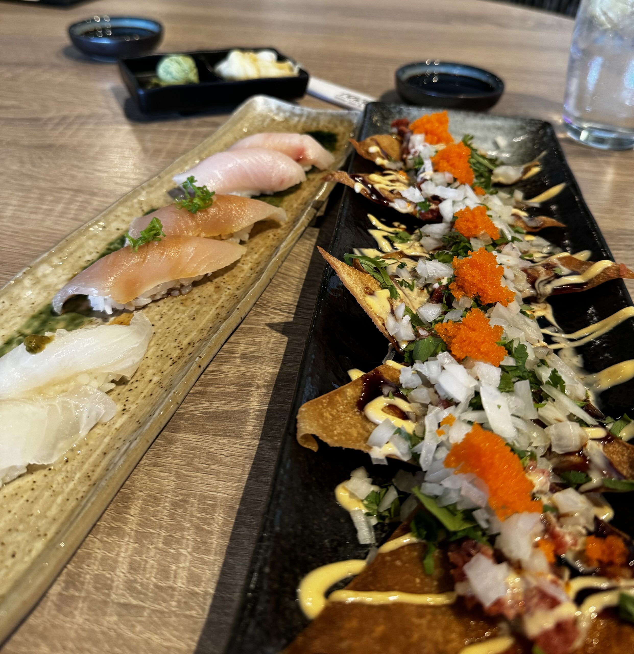 A close-up of two stylish plates at Yuzu Sushi Costa Mesa: one with assorted sushi pieces, and another with crispy nachos topped with diced onions, herbs, sauces, and bright orange roe. Bowls of soy sauce and pickled ginger are in the background.