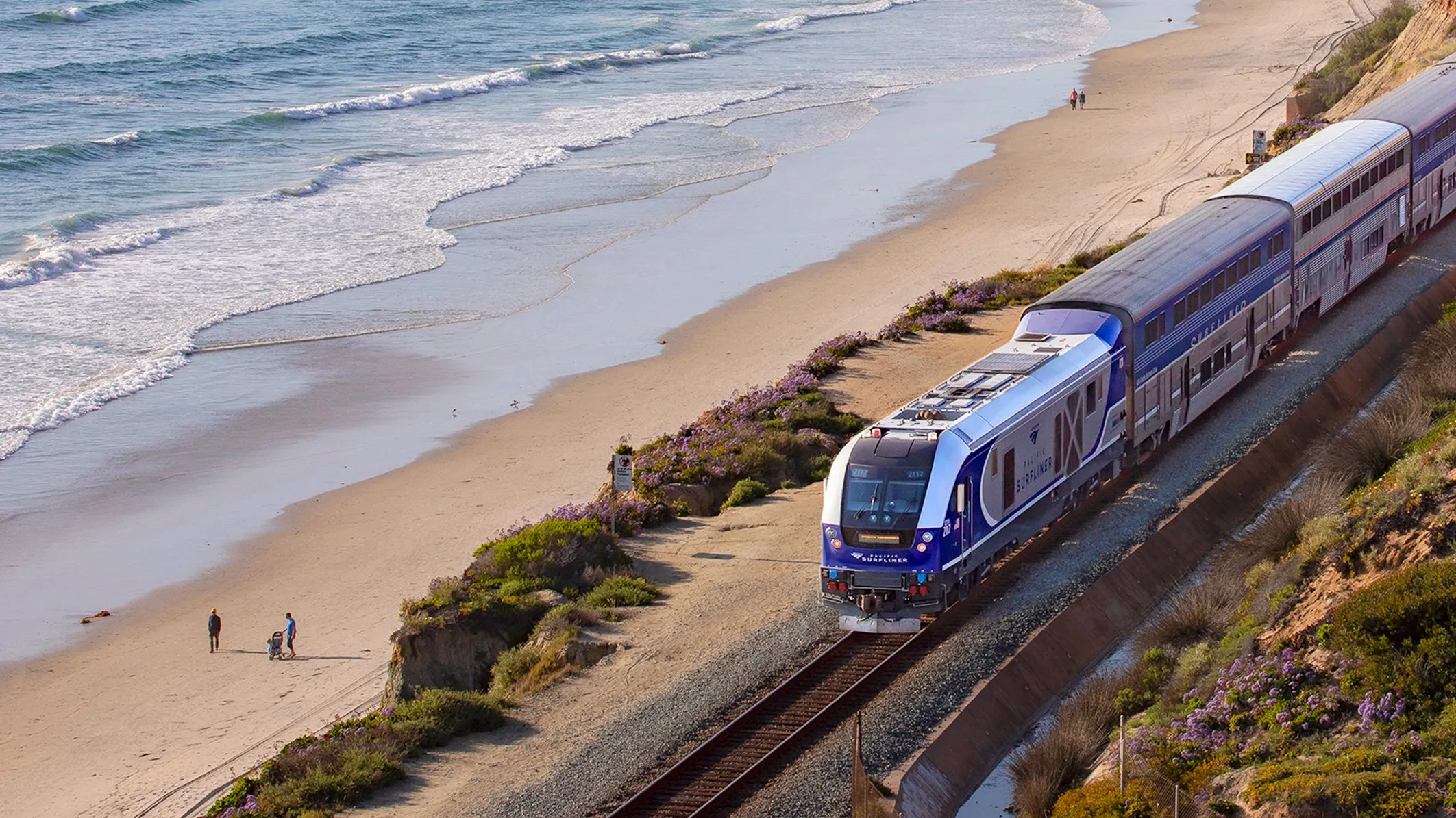 A modern blue and silver train, ideal for getting to Costa Mesa from the airport, travels along tracks beside a sandy beach with waves, while a few people walk on the shore nearby under a partly cloudy sky.