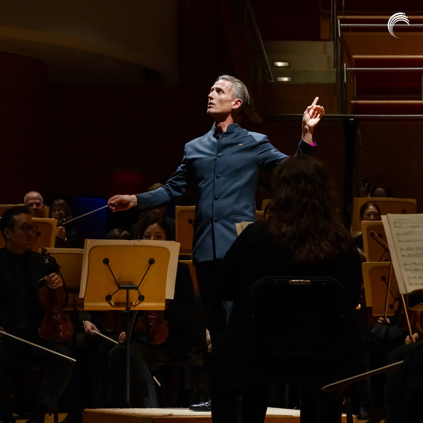 A conductor in a dark suit stands on a podium, leading an orchestra during a performance at the Segerstrom Performing Arts Center in Costa Mesa. Musicians with instruments and music stands surround him as he gestures expressively.
