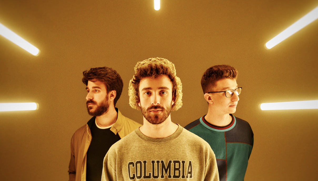 Three young men stand in front of a yellow background with glowing light beams radiating outward, as if ready for costa mesa events. The center man wears a COLUMBIA sweatshirt and furry hat, while the others sport casual shirts and glasses.