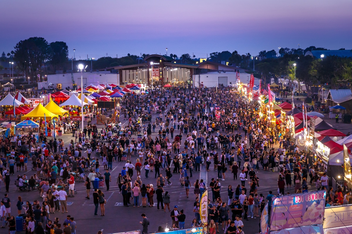 A large crowd gathers at an outdoor fair at dusk, surrounded by colorful tents, bright lights, and food stalls. People walk, eat, and socialize under the evening sky—a lively scene typical of Costa Mesa events.
