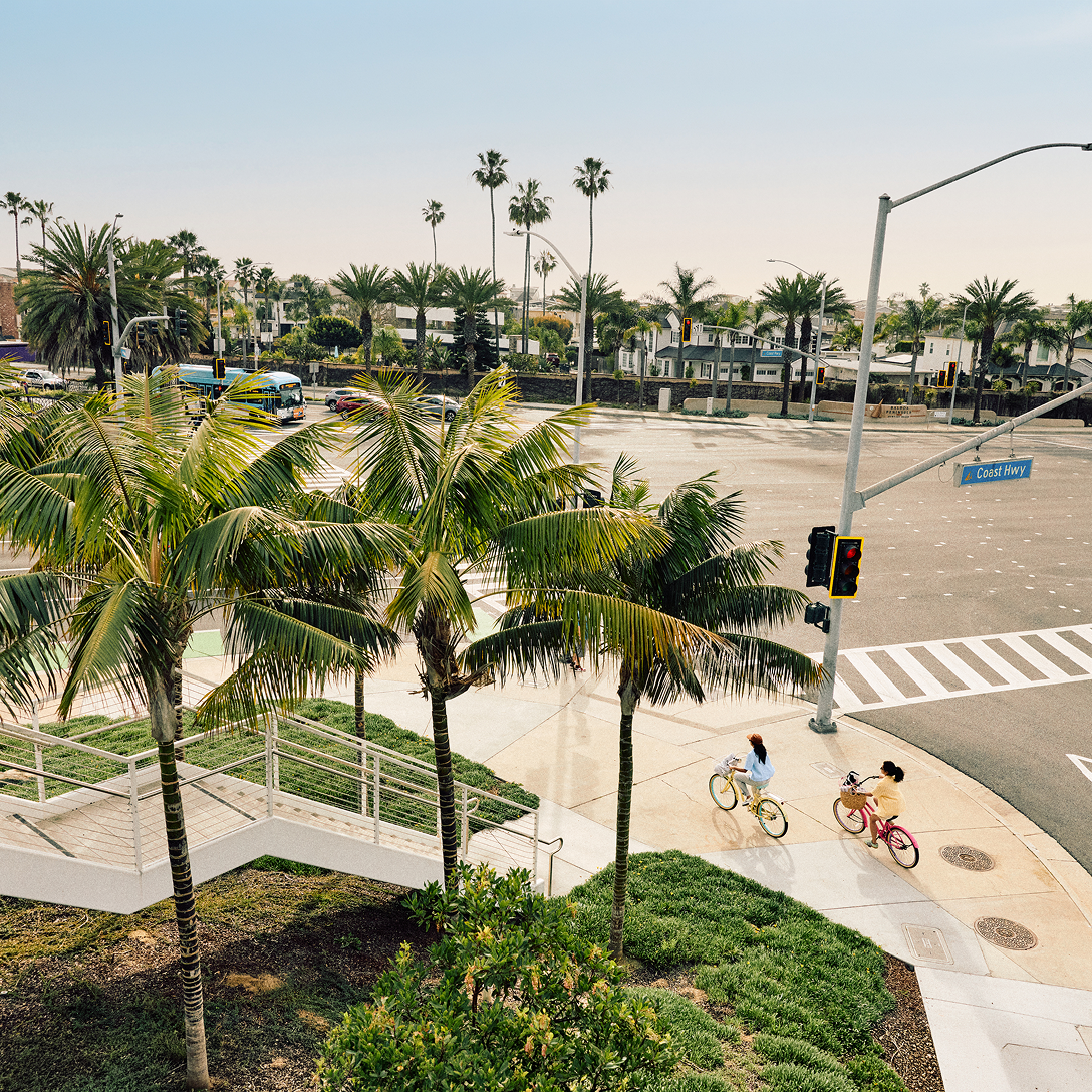 Two people ride bikes on a palm tree-lined sidewalk near a wide intersection in Costa Mesa, with crosswalks and street signs visible under a clear sky. It’s a sunny, peaceful day in this vibrant Southern California city.