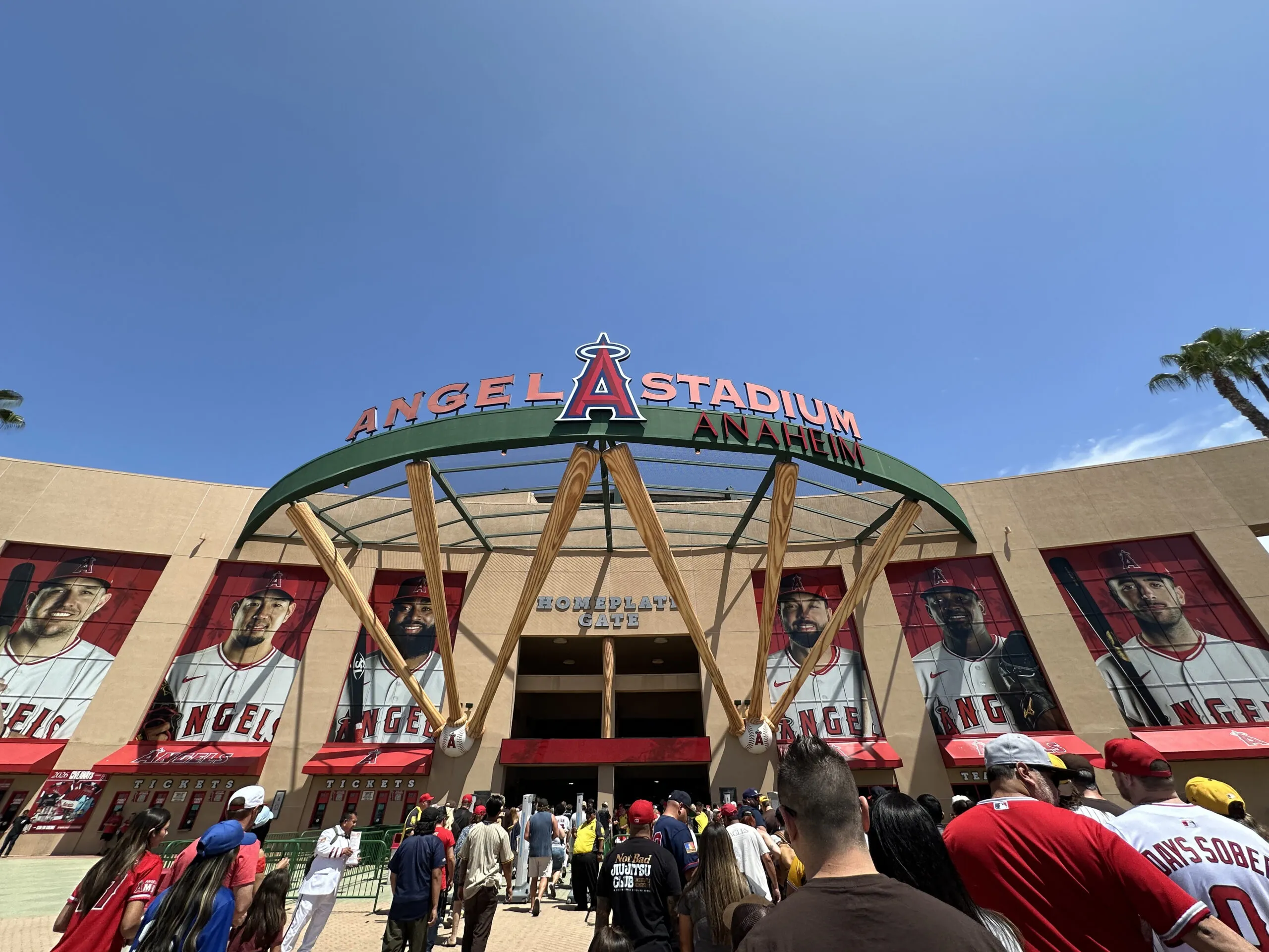 Crowds gather outside Angel Stadium of Anaheim under a sunny blue sky, with large player banners and the red “ANGEL STADIUM ANAHEIM” sign above the entrance—a popular stop for day trips from Costa Mesa.