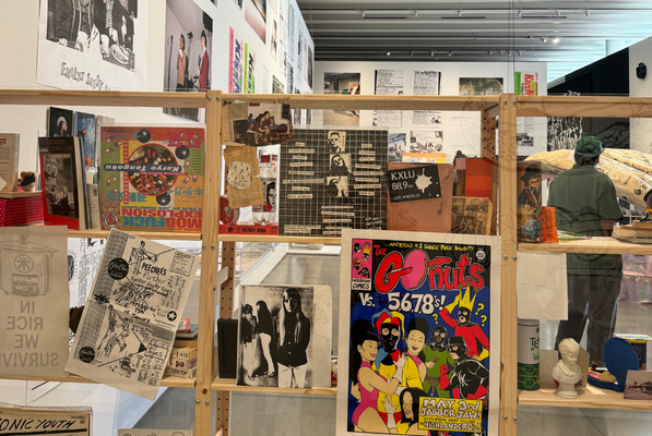 A museum exhibit displays colorful posters, zines, and black-and-white photos on a wooden frame, with a mannequin in the background—an edgy showcase of punk youth culture and one of the unique things to do in Costa Mesa.