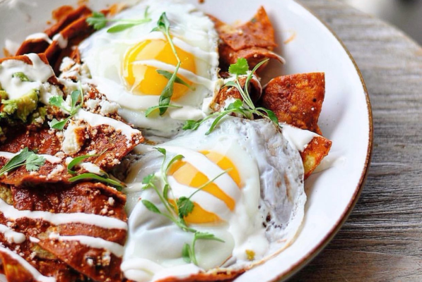 A plate of chilaquiles topped with two sunny-side-up eggs, fresh herbs, cheese, and a drizzle of crema, served on a white dish on a wooden table—perfect for dining in Costa Mesa CA.