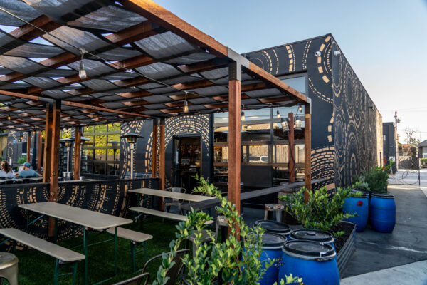 Outdoor seating area with picnic tables under a wooden pergola, next to a modern black building decorated with circular mural patterns; large blue barrels and green plants are in the foreground.