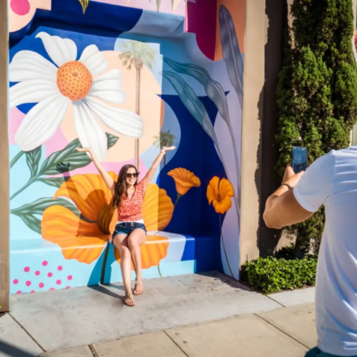 A woman in sunglasses poses with arms raised in front of a colorful mural featuring large flowers, capturing the vibrant spirit you’ll find when you visit Costa Mesa, as someone snaps her photo on a sunny sidewalk.