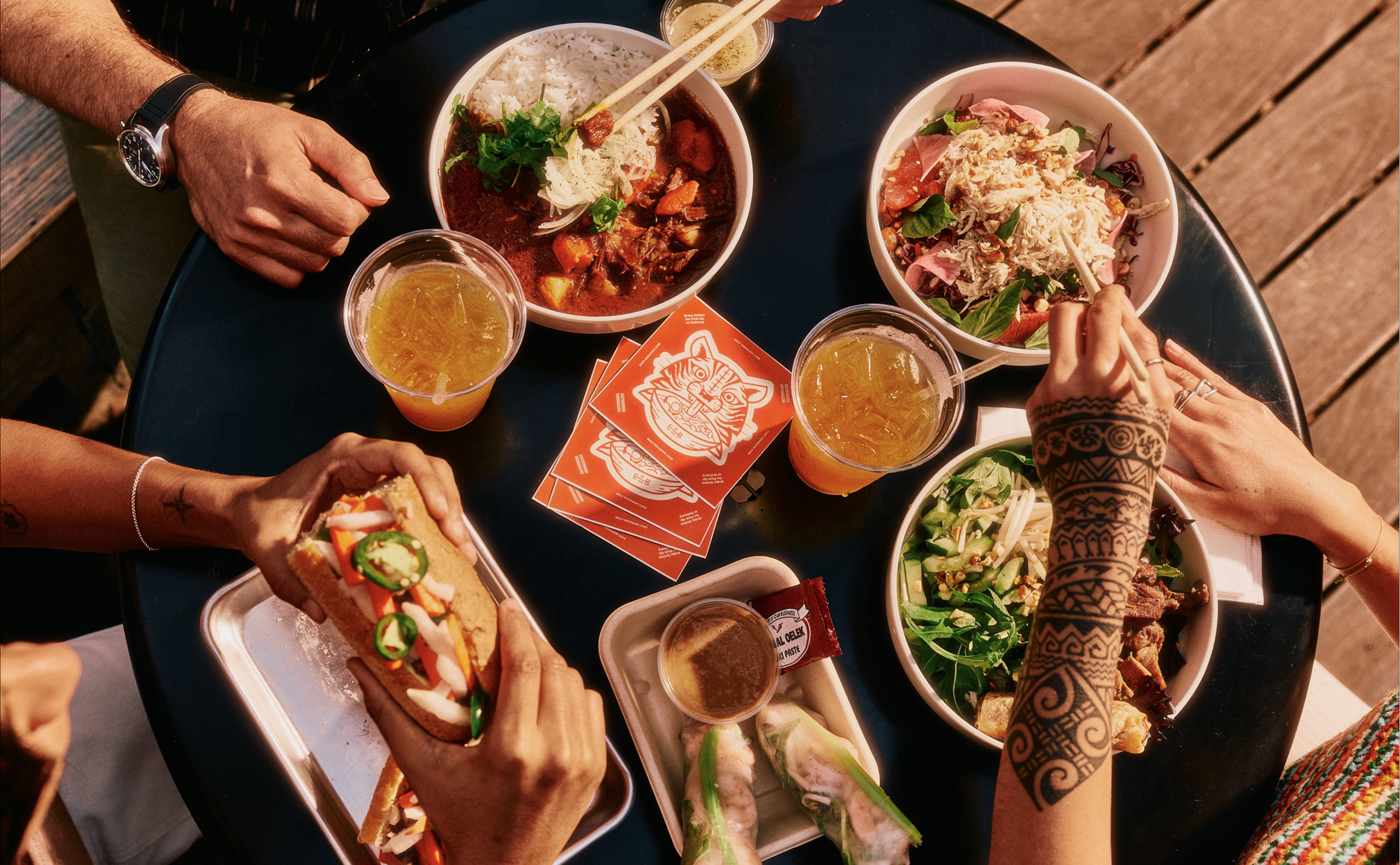 Four people enjoy Asian-inspired dishes and drinks at a round black table while dining in Costa Mesa, CA. The scene features rice bowls, salad, a sandwich, spring rolls, iced beverages, henna-adorned hands, and orange cards with a fox logo.