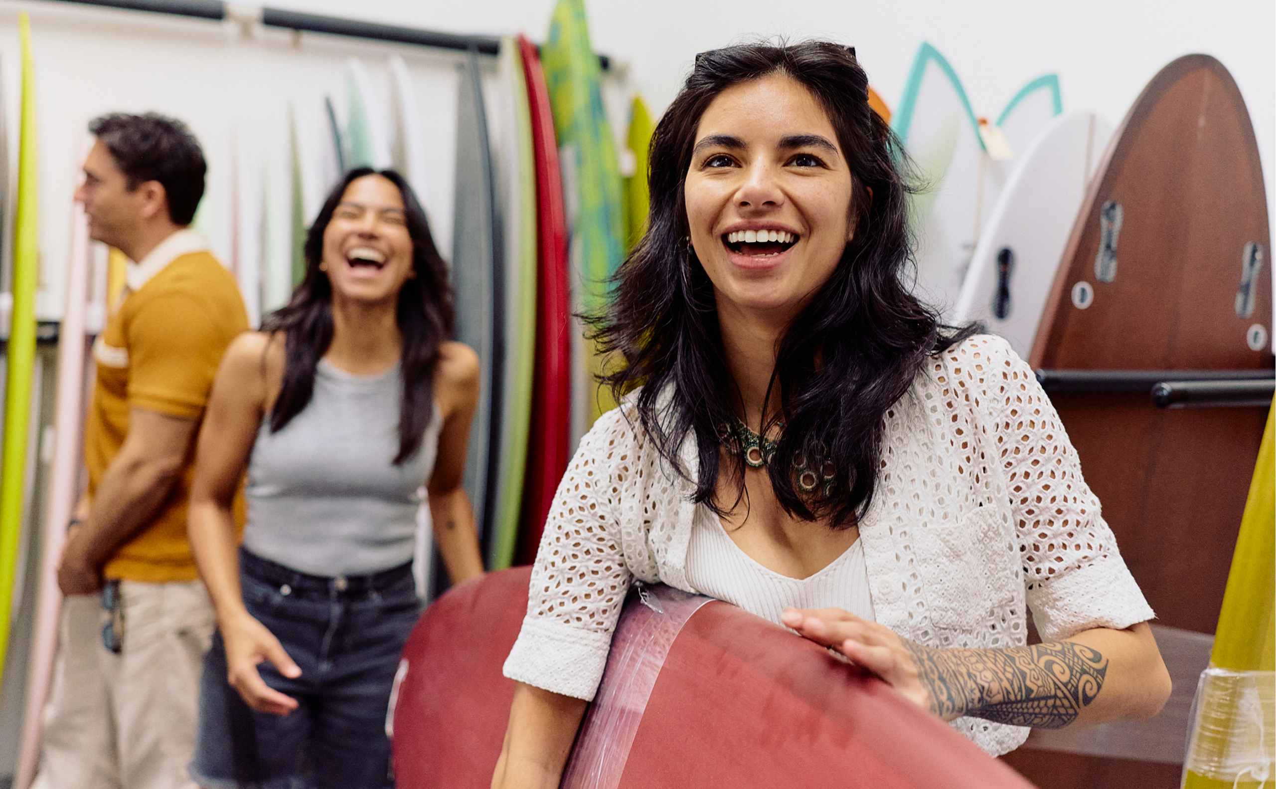 Three people stand in a surf shop, smiling and laughing among colorful surfboards. A woman in front holds a red board, while a man and another woman interact in the background—capturing the cheerful vibe of Costa Mesa events.