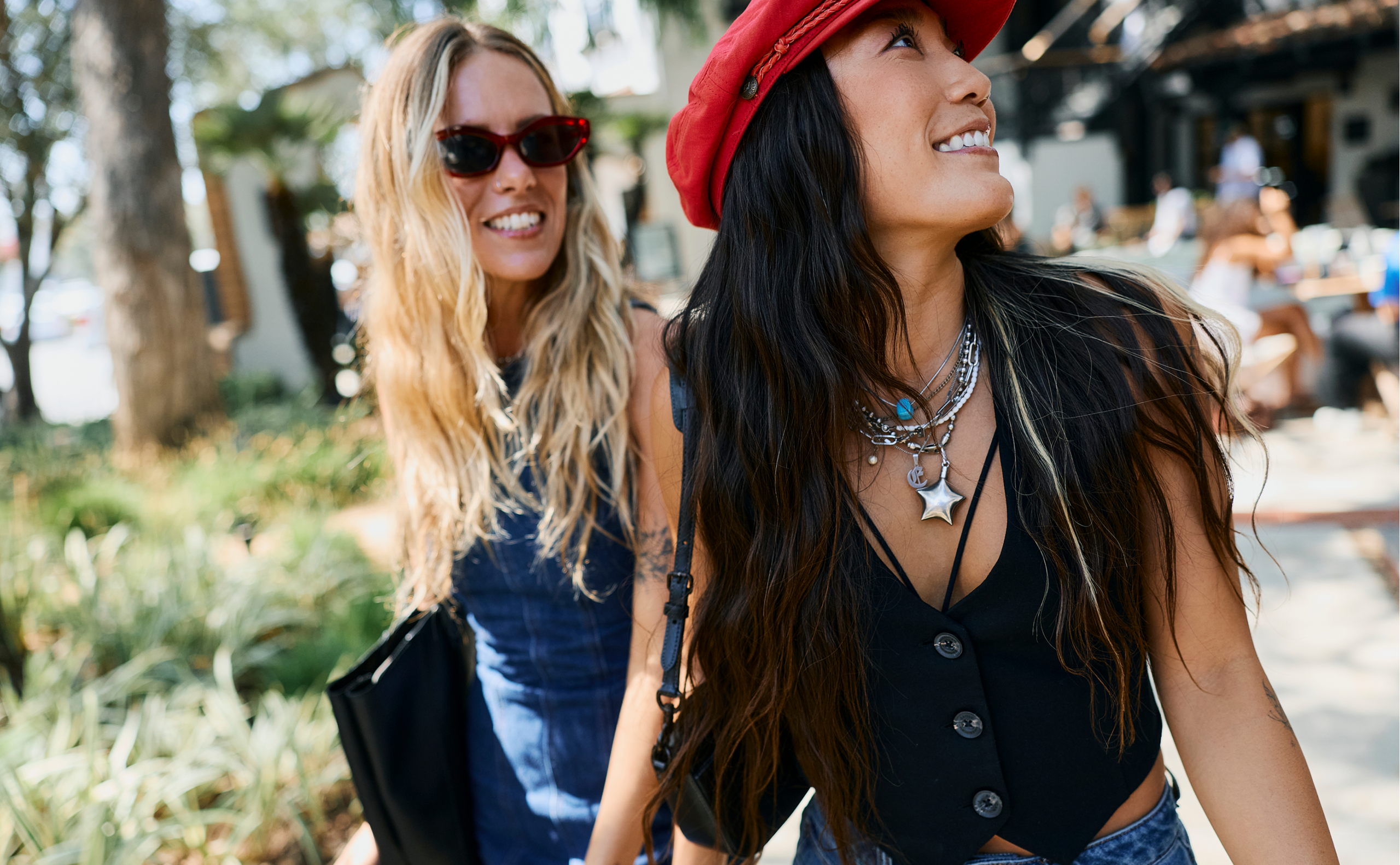 Two women walking outdoors, smiling and enjoying a sunny day exploring Costa Mesa attractions. One wears a red hat and layered necklaces, while the other has sunglasses and long blonde hair. Greenery and people in the background.