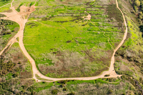 Aerial view of a large grassy hill with winding dirt trails, surrounded by green and brown vegetation. Residential houses border the edge in the background, just a short drive from popular beaches near Costa Mesa.