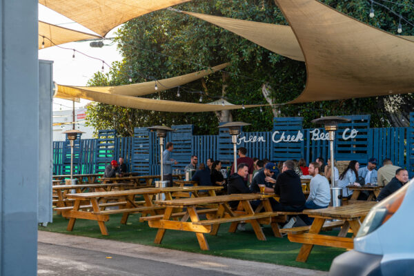 People are sitting at wooden picnic tables under sun shades in the outdoor seating area of a Costa Mesa brewery, with string lights and patio heaters adding warmth. A sign on the fence reads Green Cheek Beer Co.