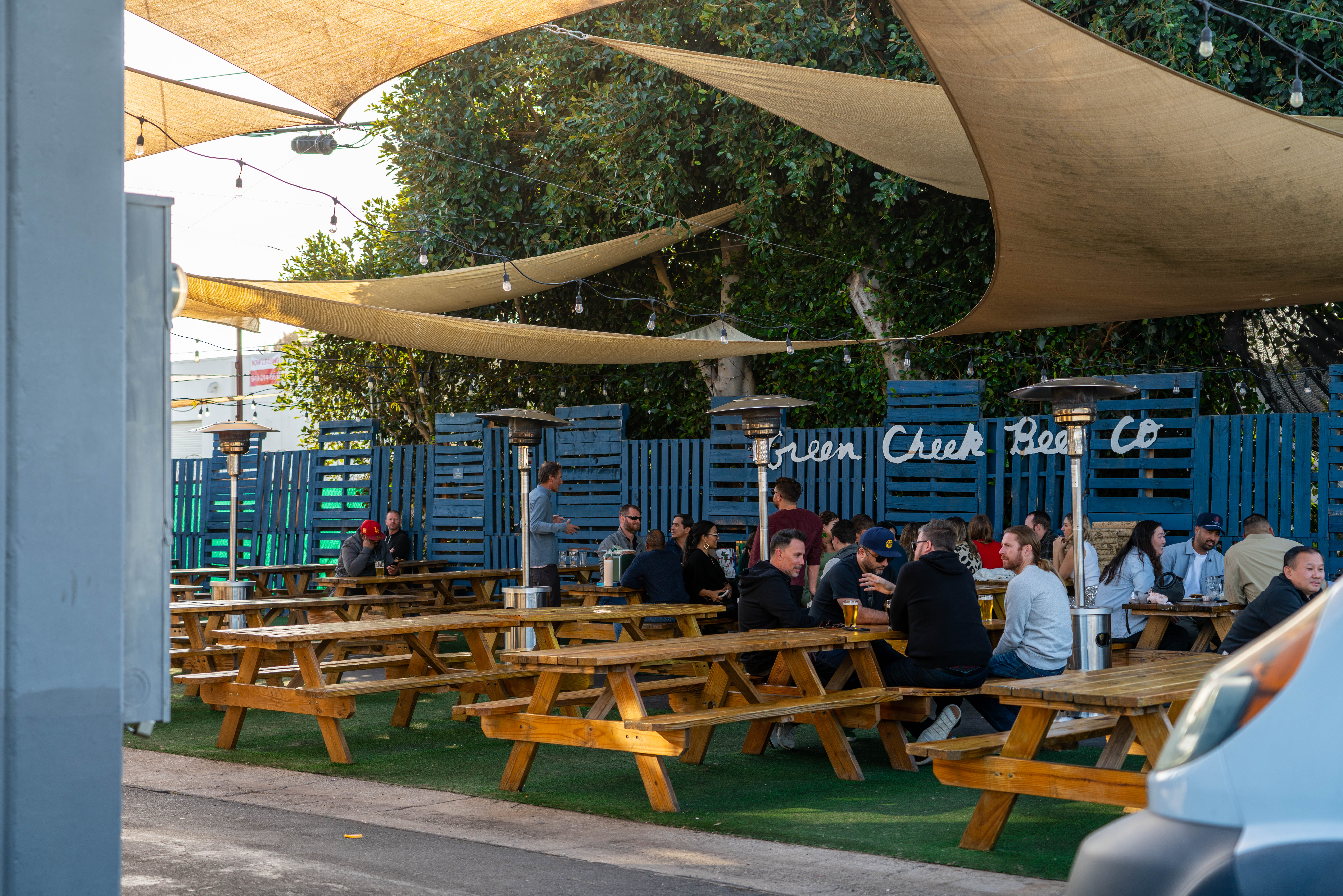 People are sitting at wooden picnic tables under sun shades in the outdoor seating area of a Costa Mesa brewery, with string lights and patio heaters adding warmth. A sign on the fence reads Green Cheek Beer Co.