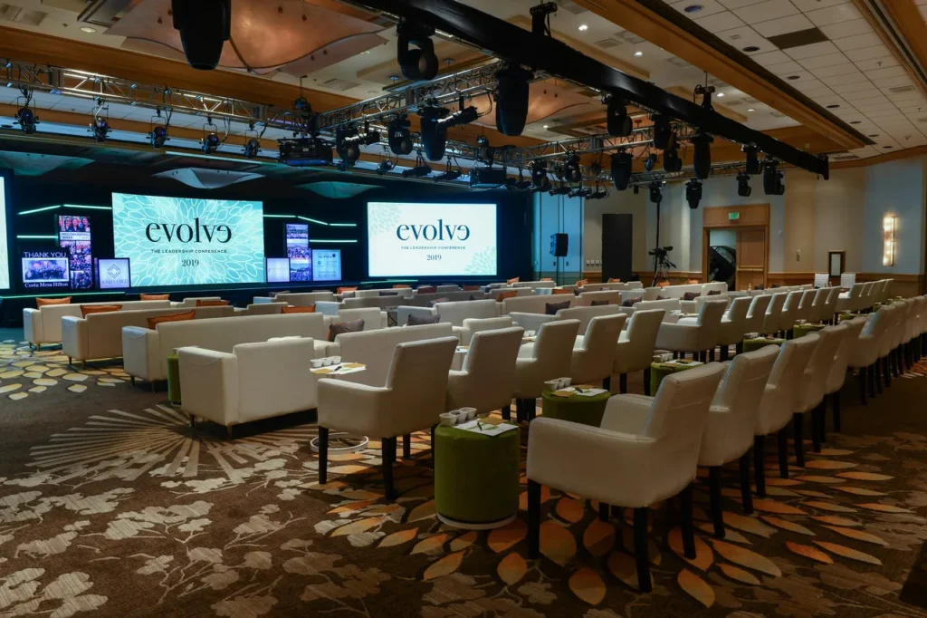 A modern conference room among top event venues in Costa Mesa, featuring white armchairs facing large digital screens displaying “evolve 2019.” The space is well-lit, with patterned carpet and stage lighting overhead.