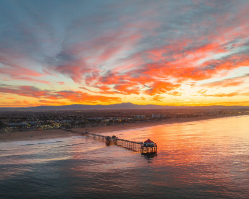 A long pier extends over calm ocean water at sunset, with vibrant orange, pink, and purple clouds filling the sky above a coastal town—capturing the beauty of beaches near Costa Mesa.