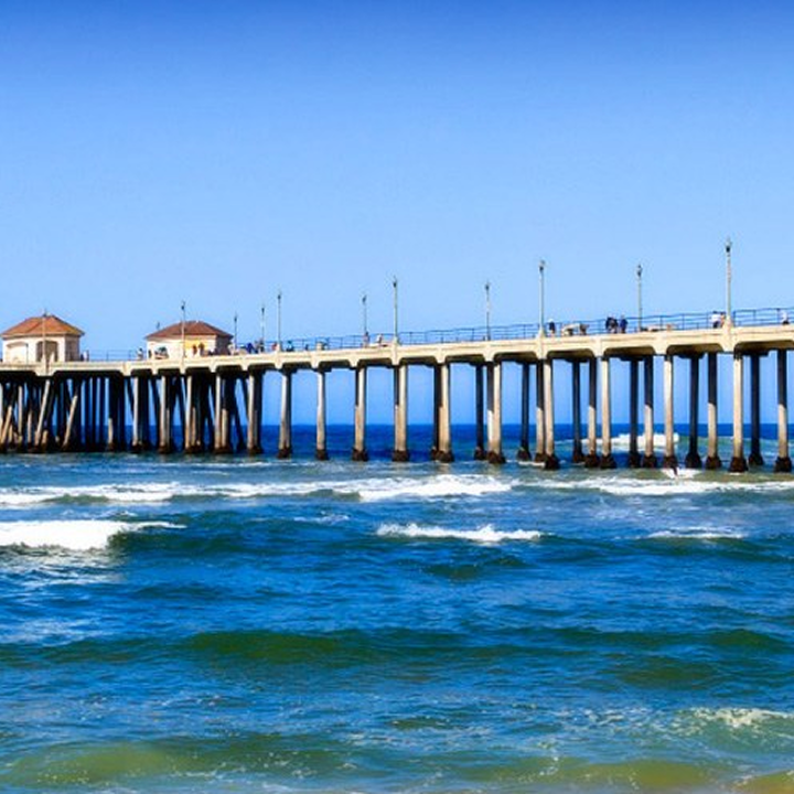 A long pier with two small buildings extends over the ocean in pet friendly Costa Mesa, with people walking along it. Waves crash below, and the sky is clear and bright blue.