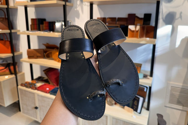 A hand holds up a pair of black leather sandals with toe loops inside a Costa Mesa store, with shelves displaying wallets and leather goods in the background.