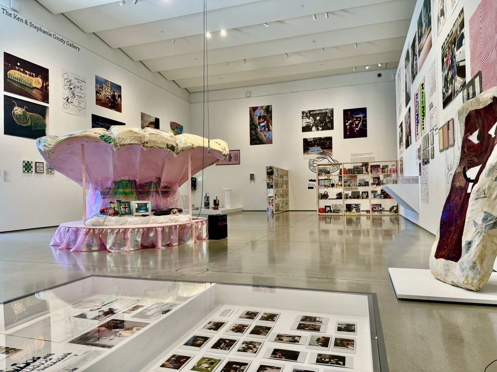 A contemporary art gallery in Costa Mesa with high ceilings displays large sculptures, colorful wall art, photos, and mixed media installations. The centerpiece is a pink carousel structure draped in fabric, surrounded by various artworks.