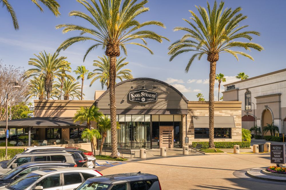 Street view of Karl Strauss Brewing Company, a popular Costa Mesa brewery, surrounded by tall palm trees, with several parked cars in the foreground and a clear blue sky above.