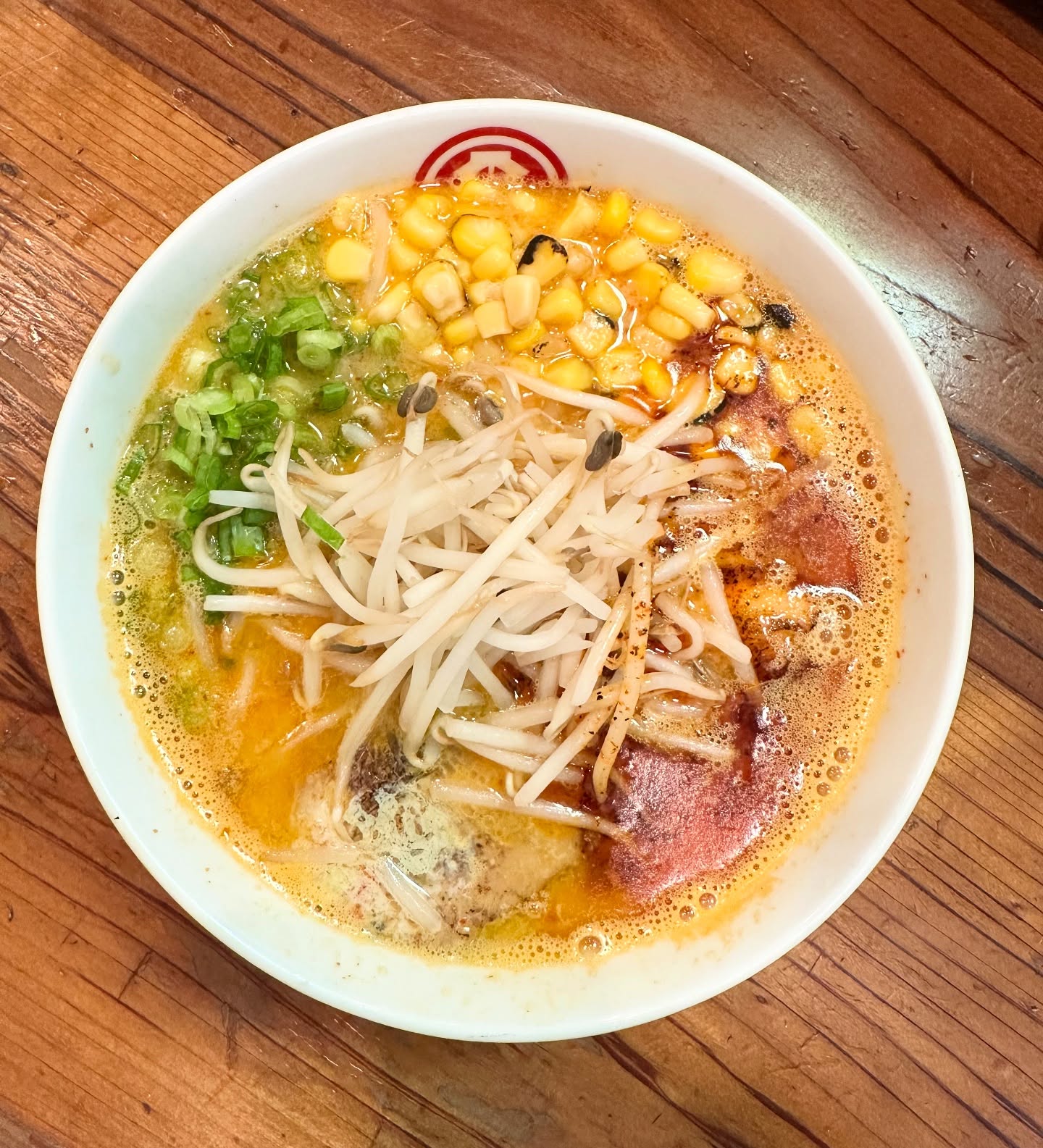 A bowl of ramen noodles Costa Mesa style, topped with corn, green onions, bean sprouts, and spices, sits on a wooden table. The rich and creamy broth fills a white bowl marked with a red logo on the rim.
