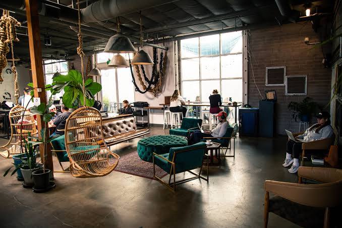 A spacious, modern café in Costa Mesa with people working on laptops, cozy armchairs, a brown leather sofa, hanging chairs, large windows flooding the coffee space with natural light, plus lush plants and stylish décor elements.