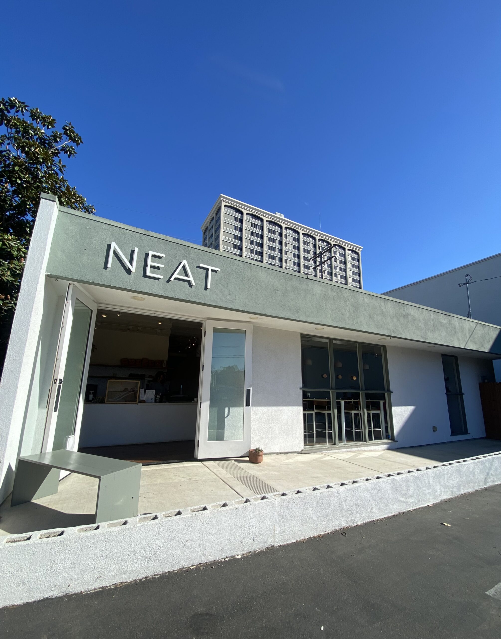 A modern café with the word NEAT displayed on its green-gray facade invites you in for coffee. Large glass doors open to a minimalist interior, while a tall building and clear blue sky set the scene in Costa Mesa.