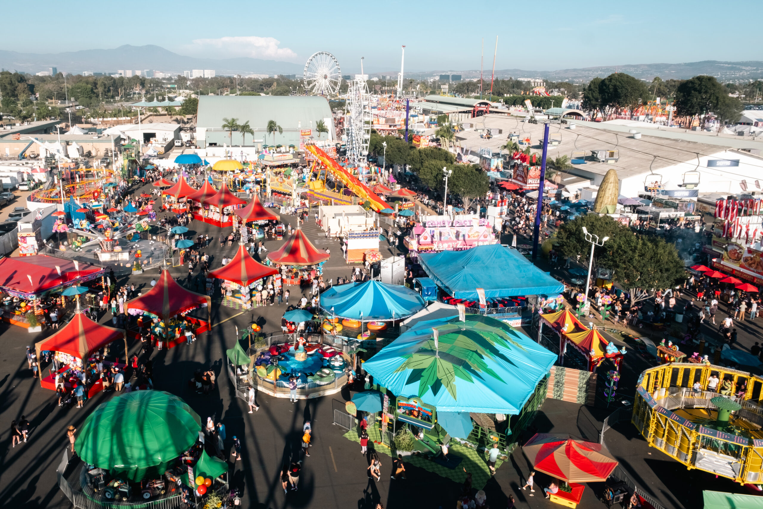 Aerial view of a busy fairground in Costa Mesa with colorful tents, rides, and attractions, including a Ferris wheel. People walk among the booths and rides under a clear blue sky with mountains visible in the distance.