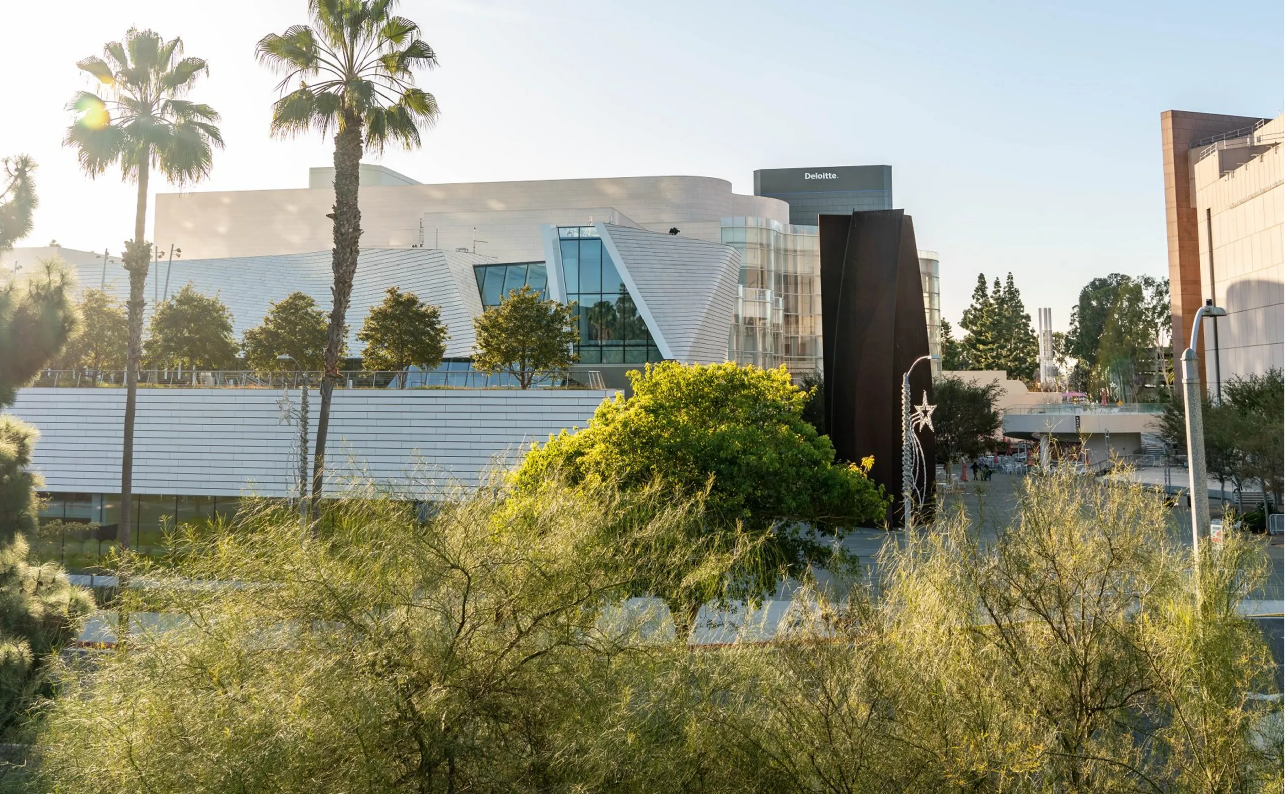 A modern building with angular roofs and large windows is surrounded by palm trees and greenery on a sunny day. A dark vertical sculpture stands in front, with a Deloitte sign visible—one of the notable Costa Mesa attractions.
