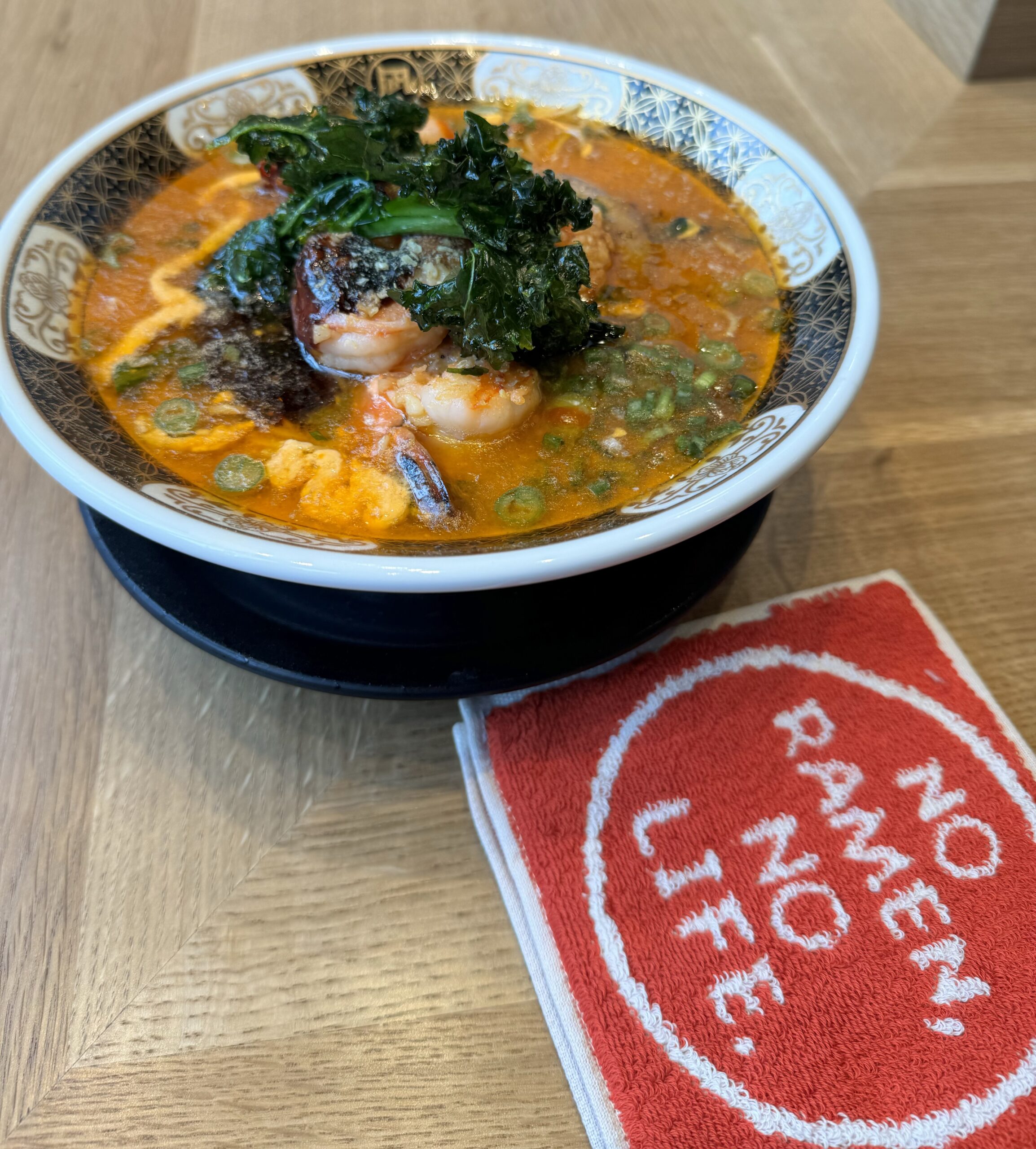 A bowl of ramen noodles Costa Mesa style, with shrimp, greens, and rich broth, sits on a wooden table beside a red napkin with white text reading, NO RAMEN, NO LIFE.