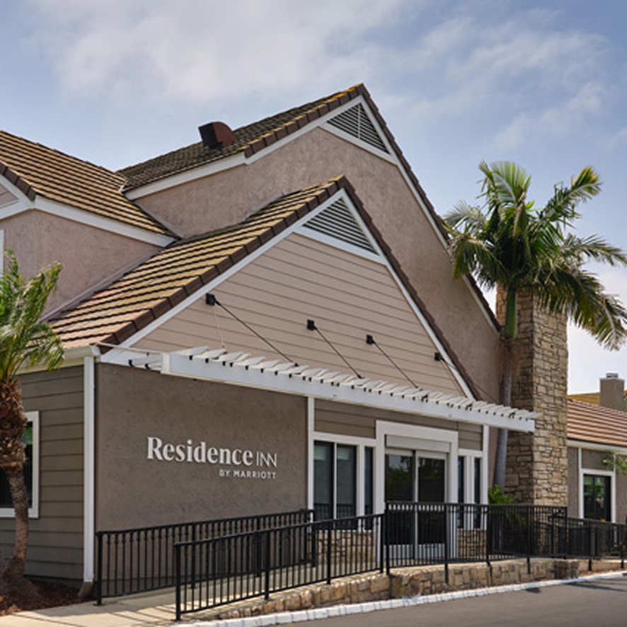 A beige, suburban-style Residence Inn by Marriott building with a sloped roof, white trim, palm trees, and a stone chimney under a partly cloudy sky—your pet friendly Costa Mesa home away from home.