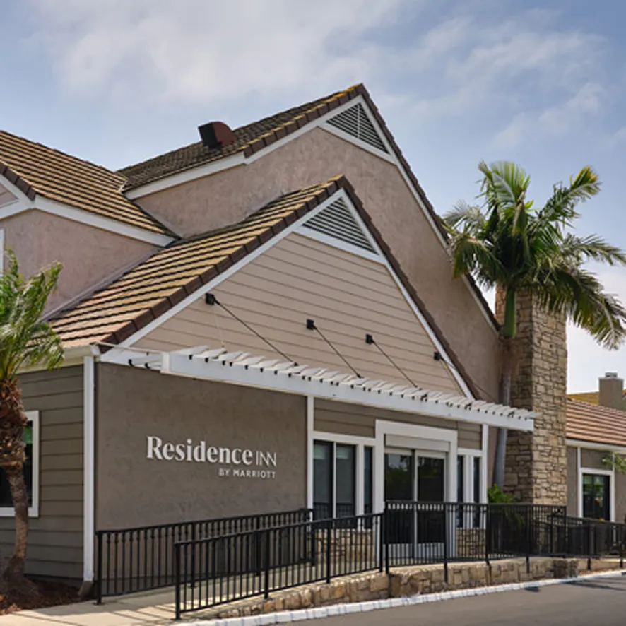 A beige, suburban-style Residence Inn by Marriott building with a sloped roof, white trim, palm trees, and a stone chimney under a partly cloudy sky—your pet friendly Costa Mesa home away from home.