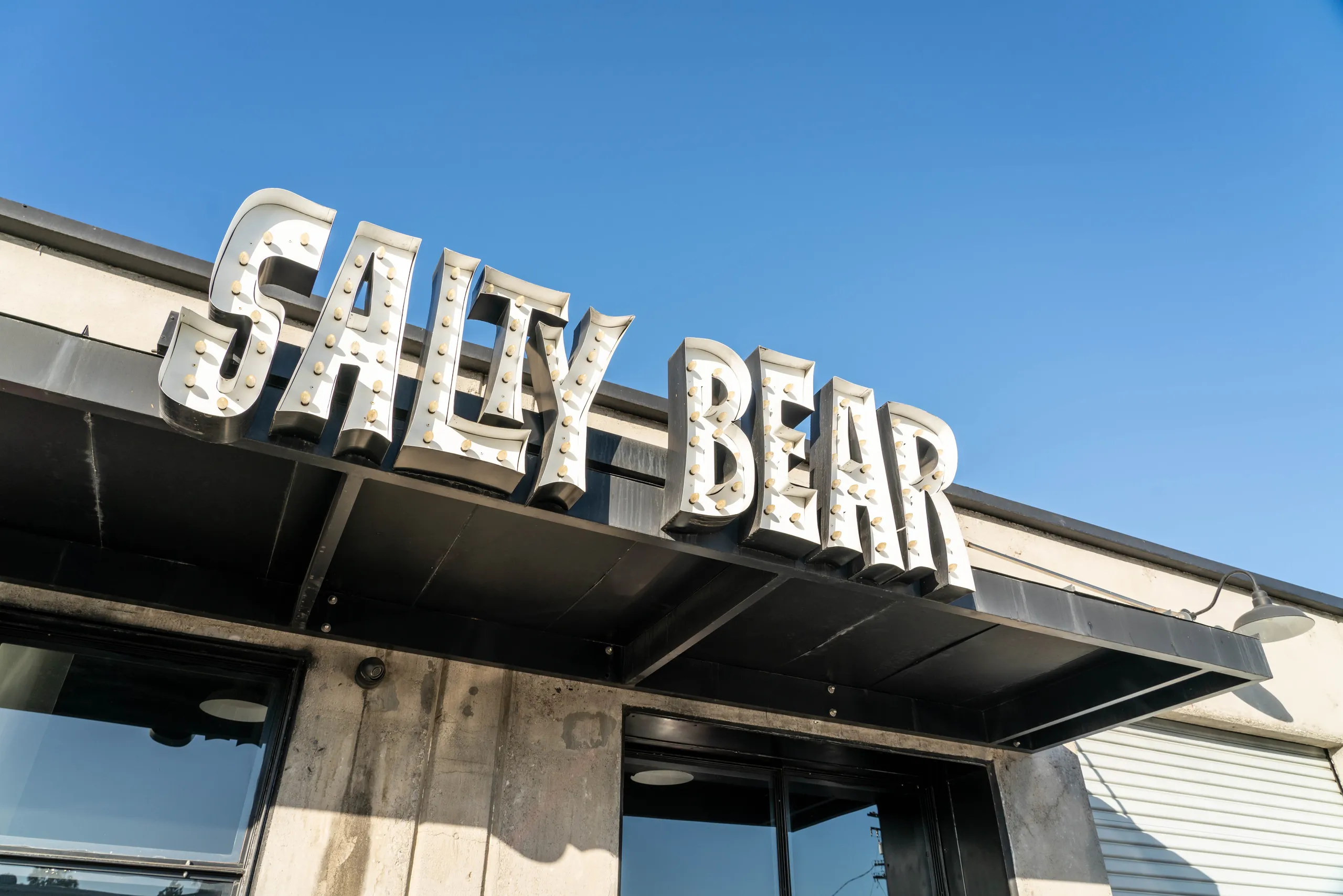 A large, retro-style sign reading SALTY BEAR is mounted above the entrance of a modern concrete building beneath a clear blue sky, marking this spot as a standout Costa Mesa brewery.
