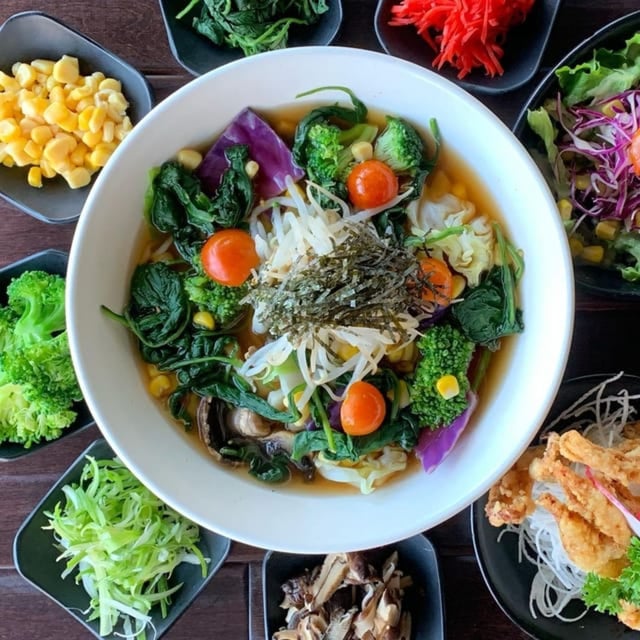 A bowl of colorful ramen noodles in Costa Mesa, topped with spinach, cherry tomatoes, seaweed, and sprouts, is surrounded by small dishes of corn, greens, shredded vegetables, and fried toppings on a wooden table.