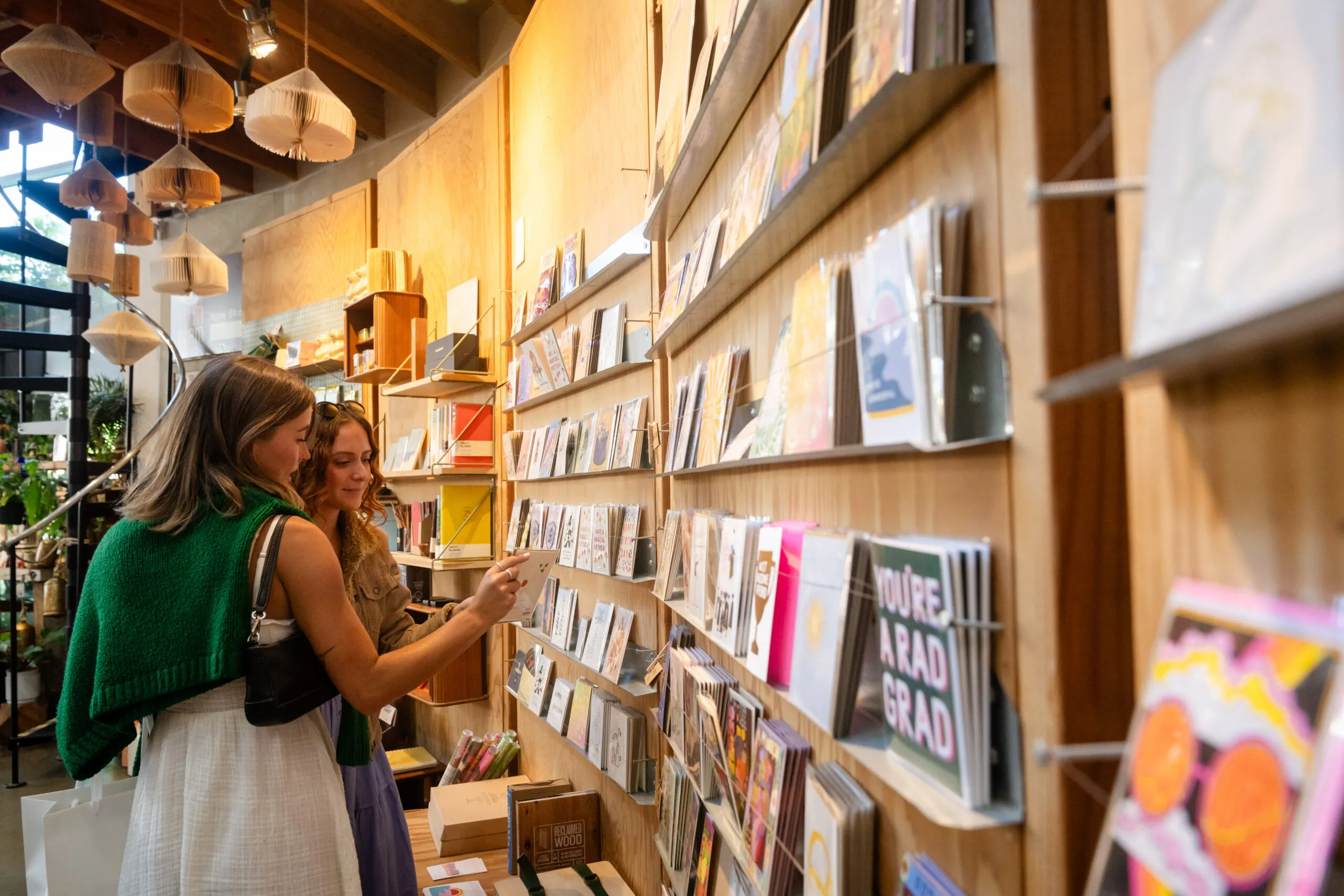 Two women browse a wall display of colorful cards and books in a cozy, well-lit shop with wooden shelves and hanging paper decorations, nestled in Costa Mesa’s vibrant theater arts district. One woman holds a card while the other looks on.