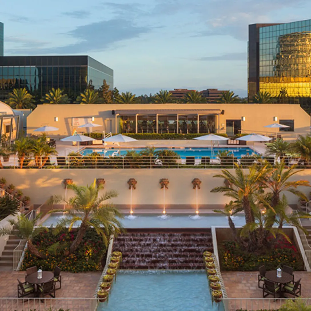 A landscaped hotel courtyard with a central water feature, palm trees, outdoor seating, and a pool area with umbrellas—set against tall glass office buildings—offers a relaxing escape near top Costa Mesa attractions under a partly cloudy sky.
