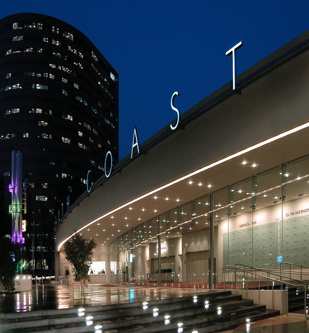 Modern glass building at night with COAST illuminated on the roof, reflecting in wet pavement; the Segerstrom Performing Arts Center Costa Mesa and a lit art sculpture are visible in the background.