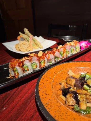 A close-up of a table at one of the best sushi Costa Mesa spots, featuring sushi rolls with fried garlic, a plate of vegetable tempura in the background, and a colorful tofu and edamame salad on an orange-rimmed plate.