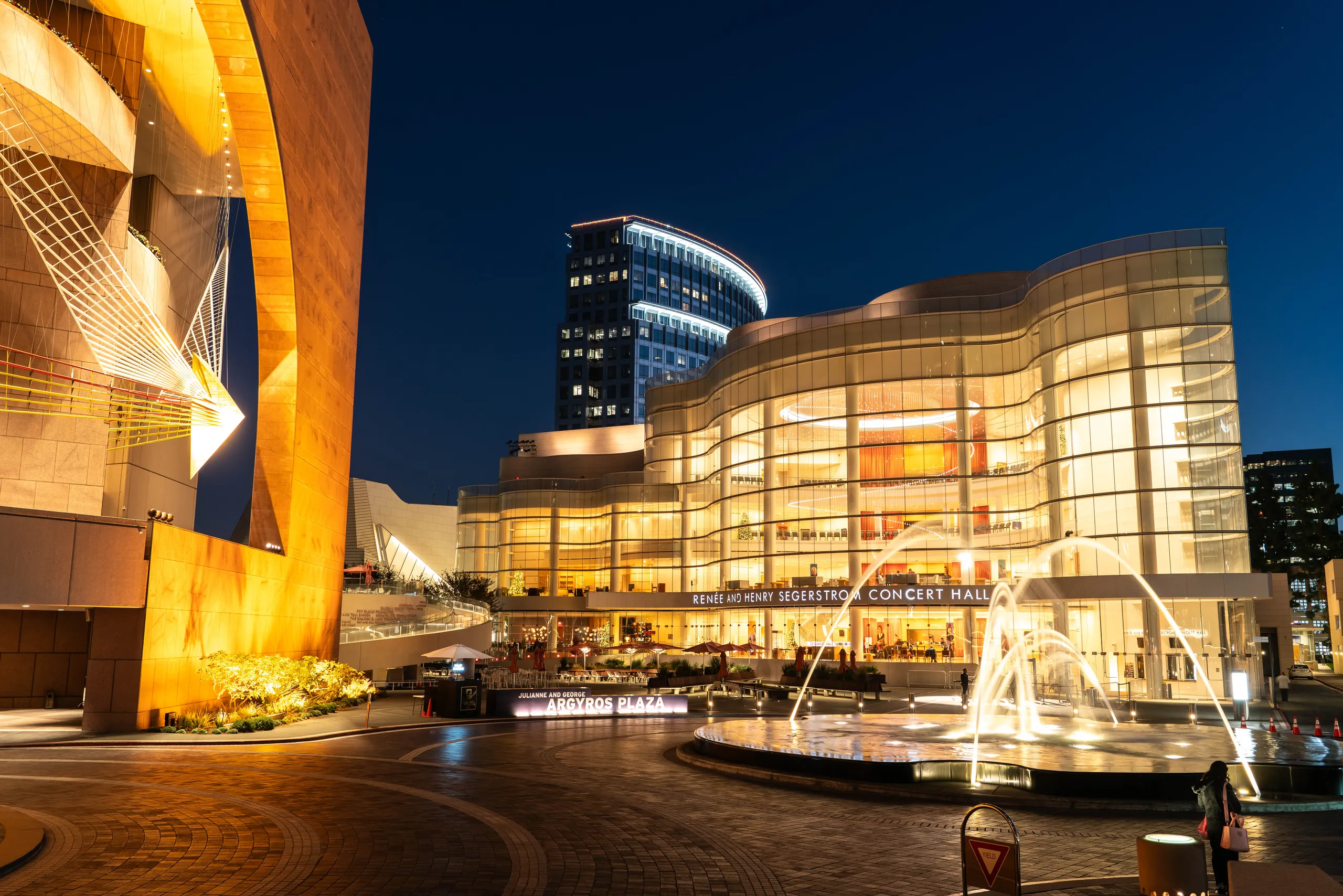 A modern concert hall with curved glass architecture and warm lighting stands next to a fountain with arcing water jets in the heart of Costa Mesa’s theater arts district, set against a city skyline at dusk.
