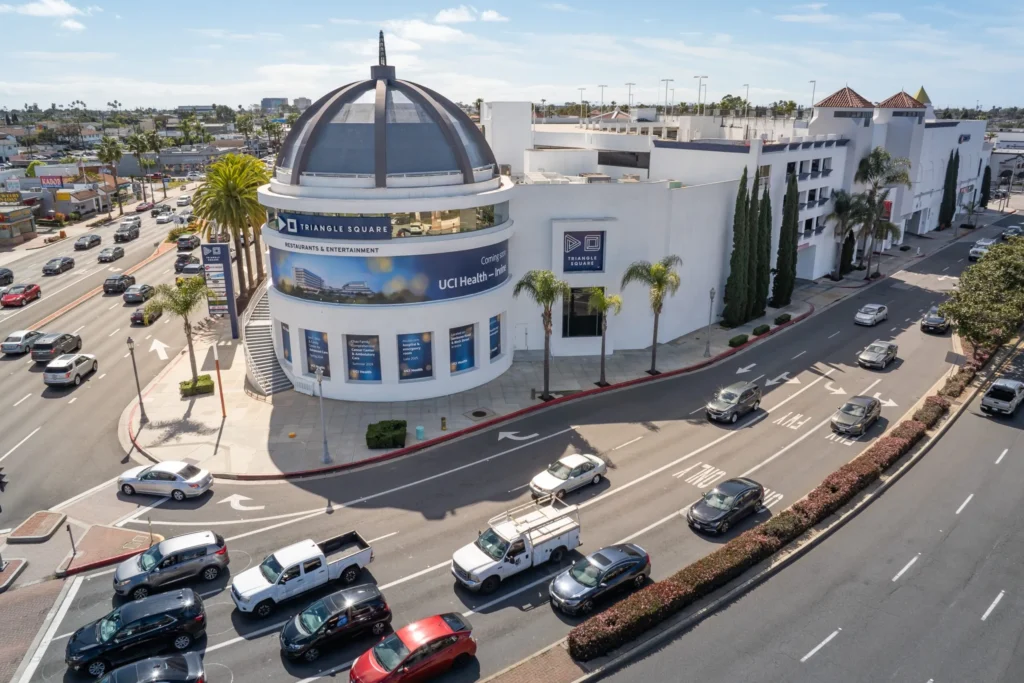 A busy urban intersection with several cars and palm trees surrounds Triangle Square, a large white shopping mall in Costa Mesa featuring a domed roof, banners, and neighboring businesses under a clear sky.