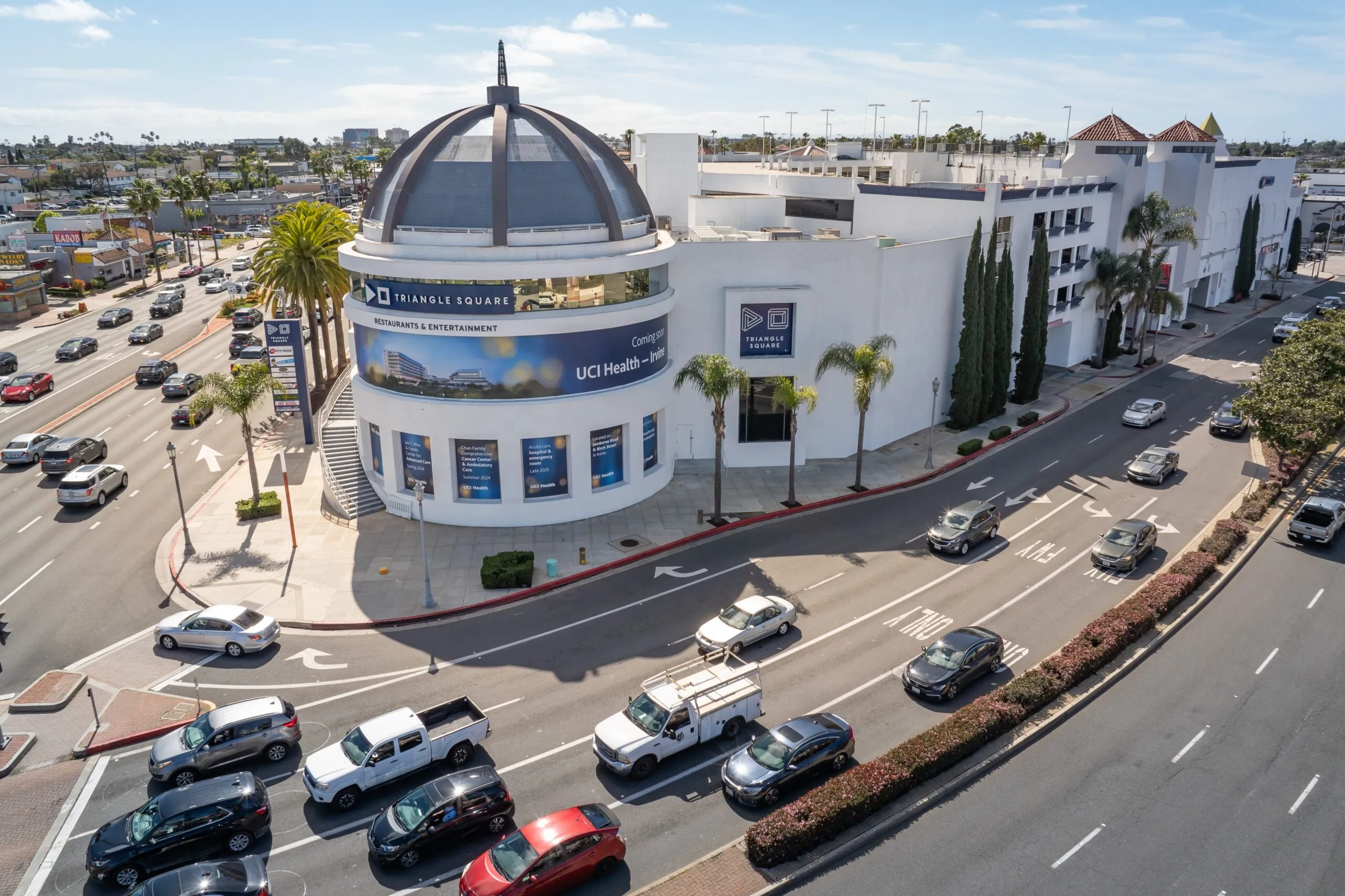 A busy urban intersection with several cars and palm trees surrounds Triangle Square, a large white shopping mall in Costa Mesa featuring a domed roof, banners, and neighboring businesses under a clear sky.