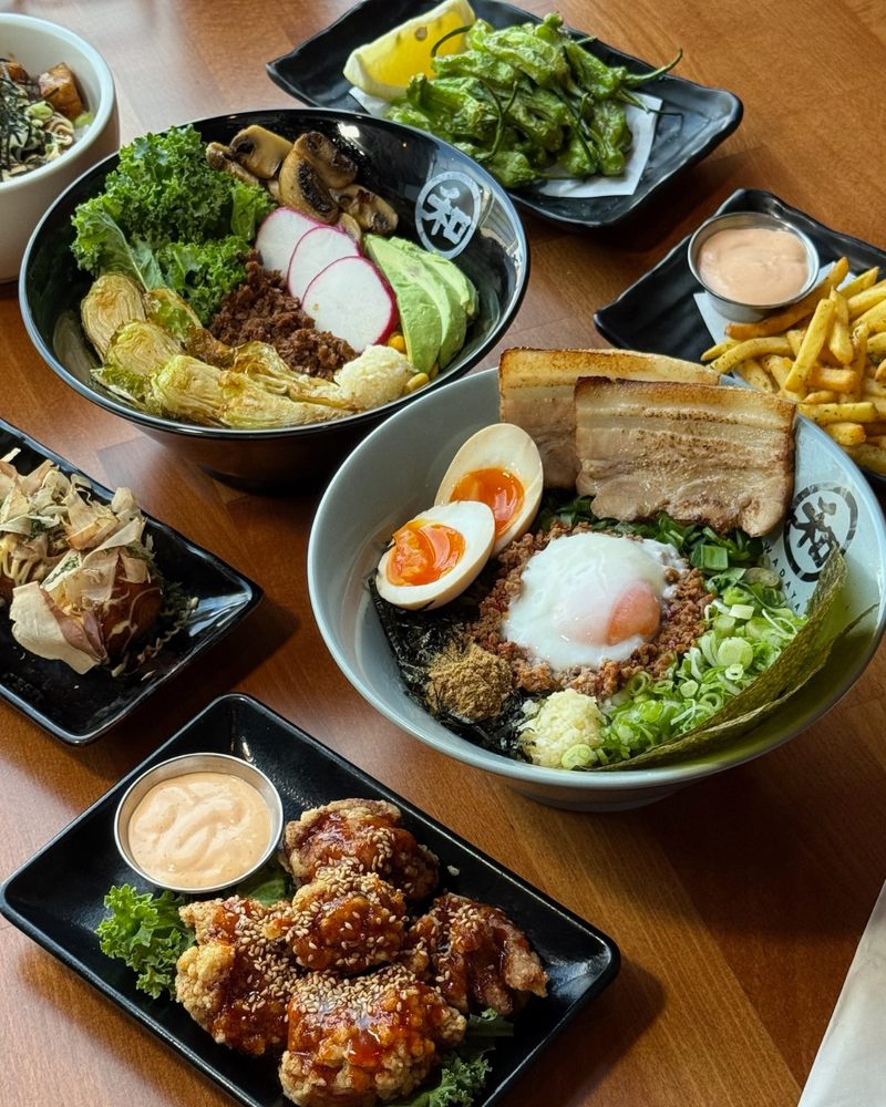 A table in Costa Mesa filled with bowls of ramen noodles topped with eggs, pork, vegetables, and seaweed, alongside plates of fried chicken, fries, shishito peppers, dipping sauces, and a grilled rice ball.