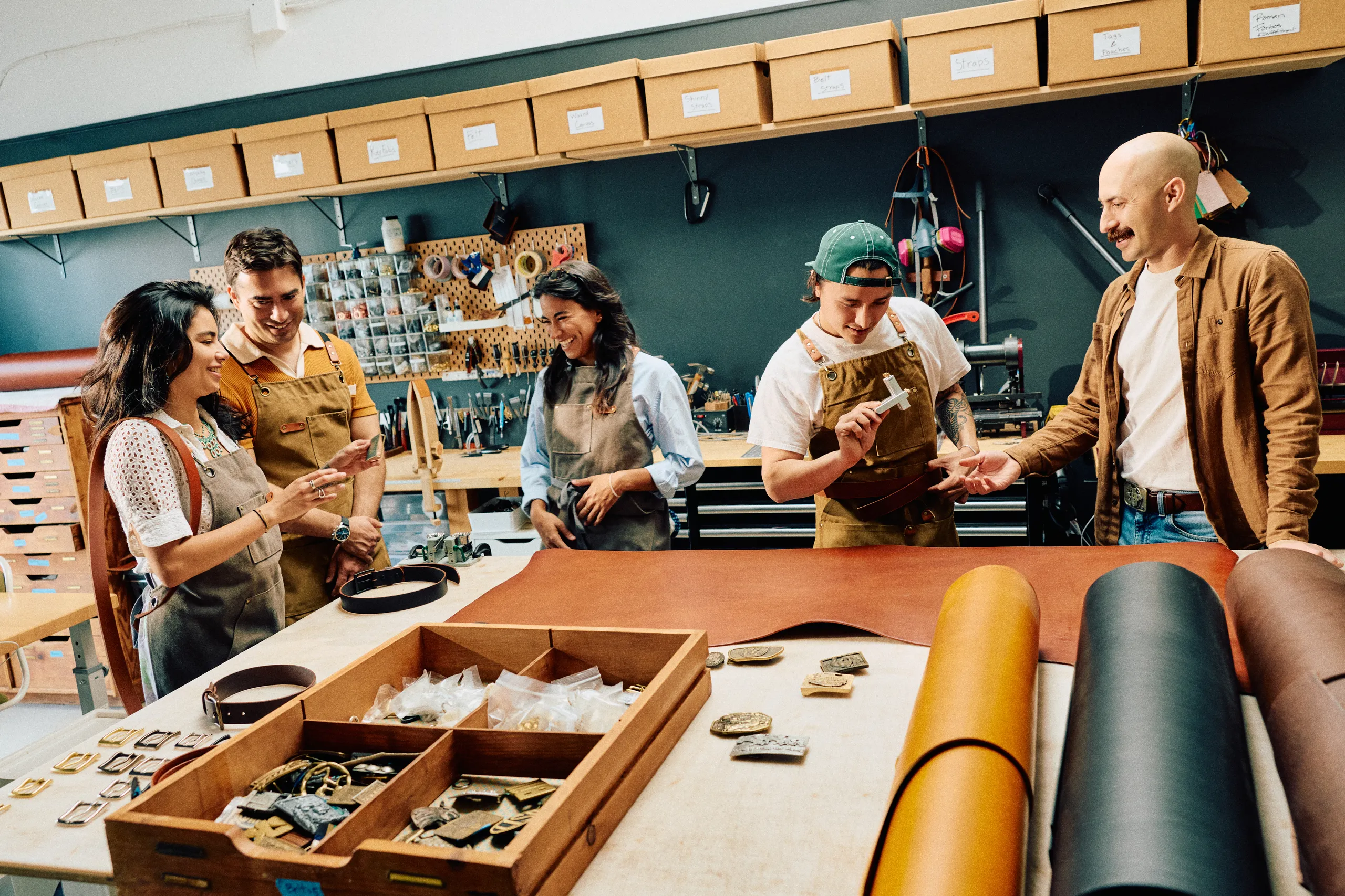 Five people gather around a worktable in a leather workshop in the heart of the theater arts district Costa Mesa, wearing aprons and smiling as they discuss leather pieces and tools, with materials and supplies organized on shelves and the table.