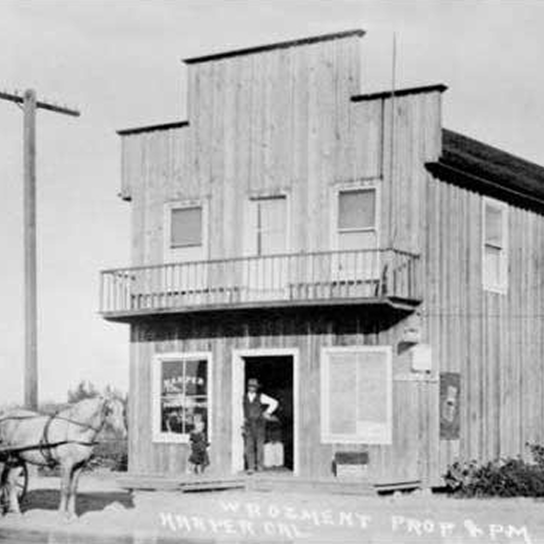 Black-and-white photo of a wooden storefront with a false front, balcony, large front windows, and a man and child at the entrance. A horse and buggy stand to the left. Text along the bottom references where is Costa Mesa.