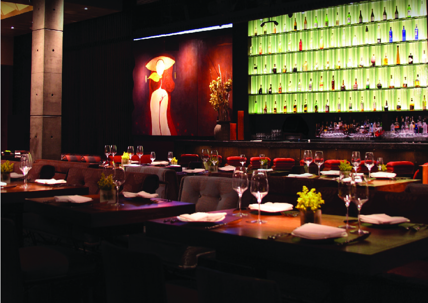 Elegant restaurant interior with neatly set tables, wine glasses, and small flower arrangements. A large painting of a woman in a white hat and a backlit wall of colorful bottles decorate the space by the bar area.
