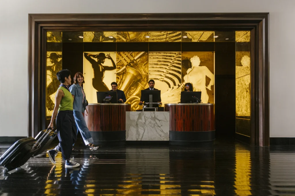 Two people with suitcases walk past a hotel front desk in Costa Mesa, California, where three staff members stand behind computers. The background features gold and black artistic panels with classical-style figures.
