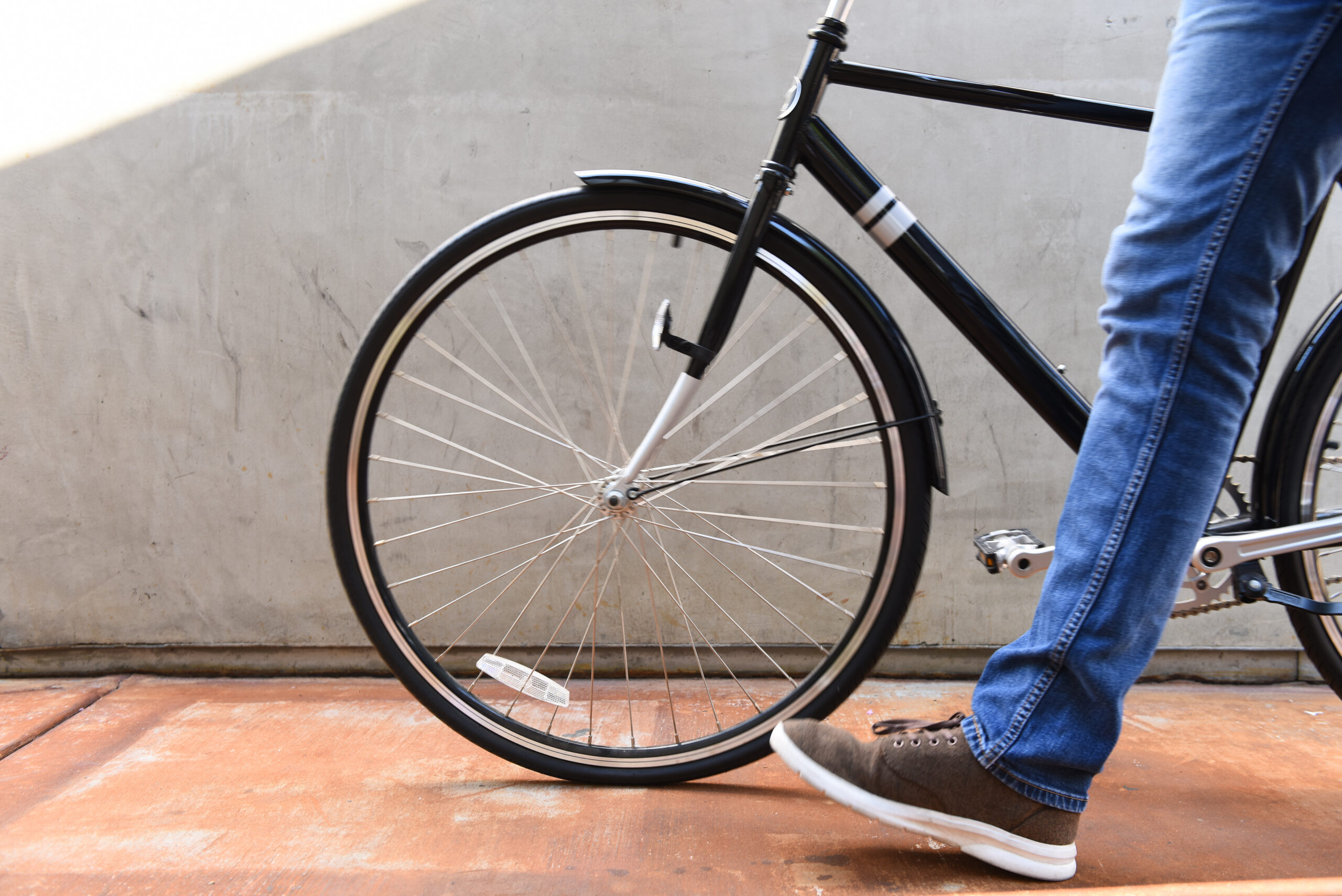 A person wearing blue jeans and brown sneakers walks alongside a black bicycle with thin tires on a brown pavement, next to a gray concrete wall, glancing at a costa mesa map in hand.