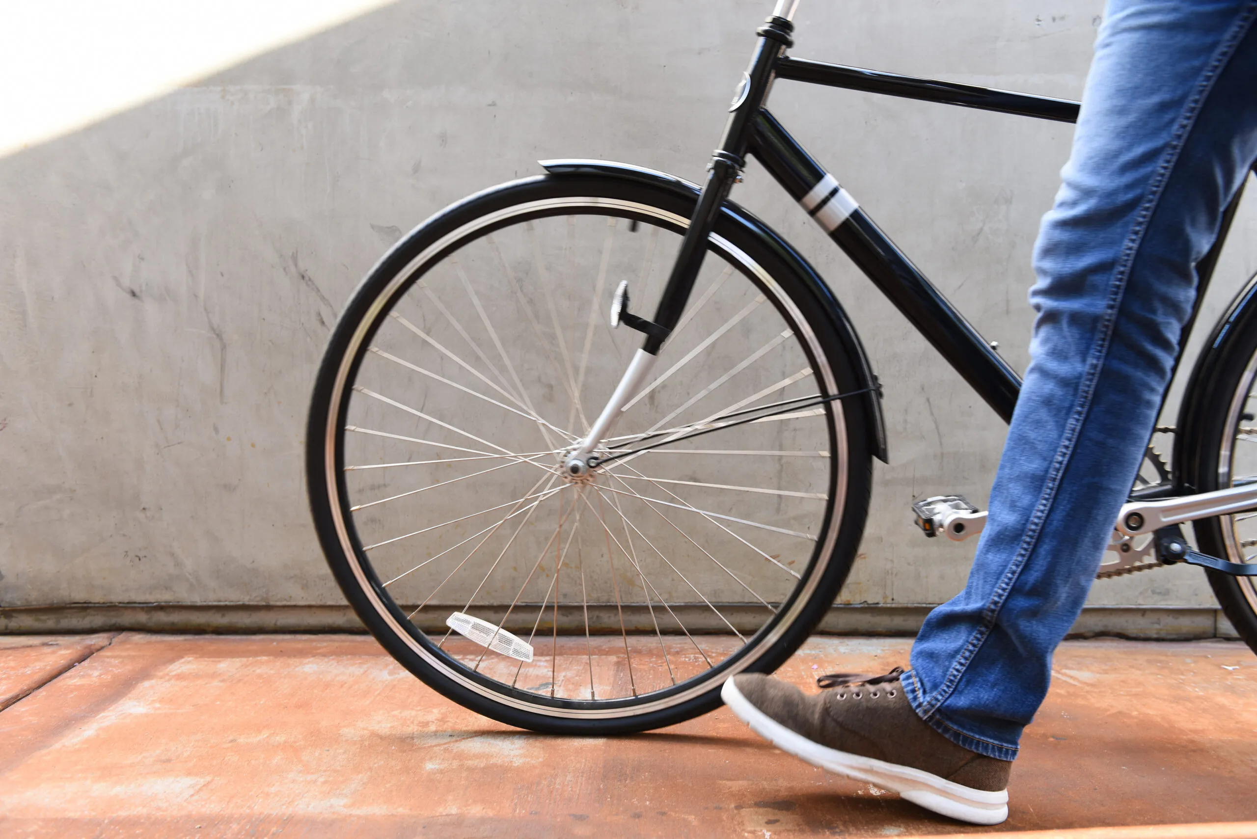 A person wearing blue jeans and brown sneakers walks alongside a black bicycle with thin tires on a brown pavement, next to a gray concrete wall, glancing at a costa mesa map in hand.