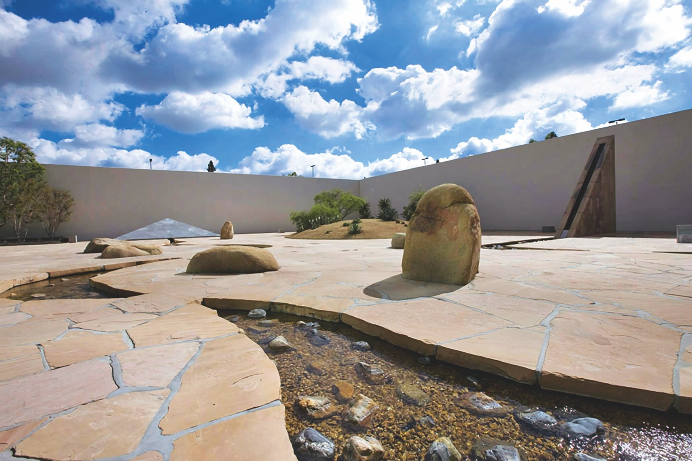 A modern outdoor courtyard in Costa Mesa, California, features large stone slabs, scattered boulders, a shallow stream, and minimal landscaping, all enclosed by high white walls under a bright sky with scattered clouds.